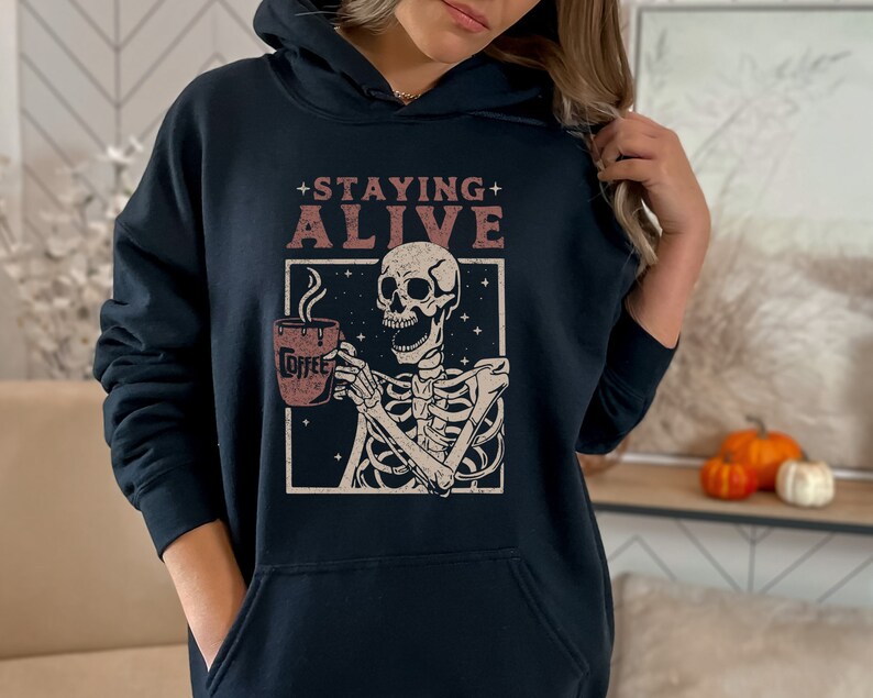 K&ouml;nnte beinhalten: Marineblauer Kapuzenpullover mit einer Grafik eines Skeletts, das eine Kaffeetasse h&auml;lt. Der Text "+ STAYING ALIVE" steht &uuml;ber dem Skelett. Auf der Kaffeetasse steht das Wort "Coffee". Das Design befindet sich in einem wei&szlig;en Rahmen mit Sternenhintergrund.