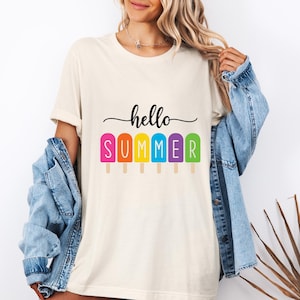 May include: Cream-colored t-shirt with the words "hello SUMMER" in black script above a row of colorful popsicle graphics. The popsicles are pink, orange, yellow, blue, and green. The shirt is worn with a blue denim jacket.