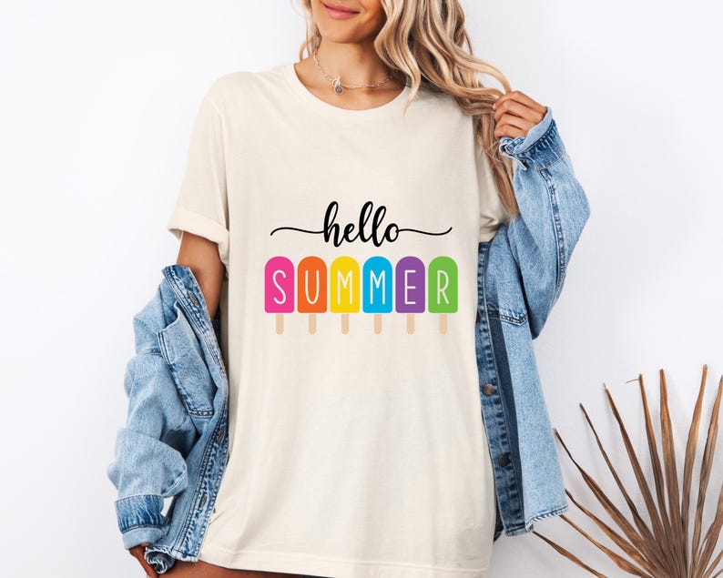 May include: Cream-colored t-shirt with the words "hello SUMMER" in black script above a row of colorful popsicle graphics. The popsicles are pink, orange, yellow, blue, and green. The shirt is worn with a blue denim jacket.