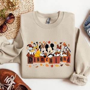 May include: A beige sweatshirt with a graphic of Mickey Mouse, Donald Duck, Goofy, and Pluto in front of a brick wall with the text "Disneyland" in brown letters. The characters are wearing fall-themed outfits and there are leaves scattered around them.