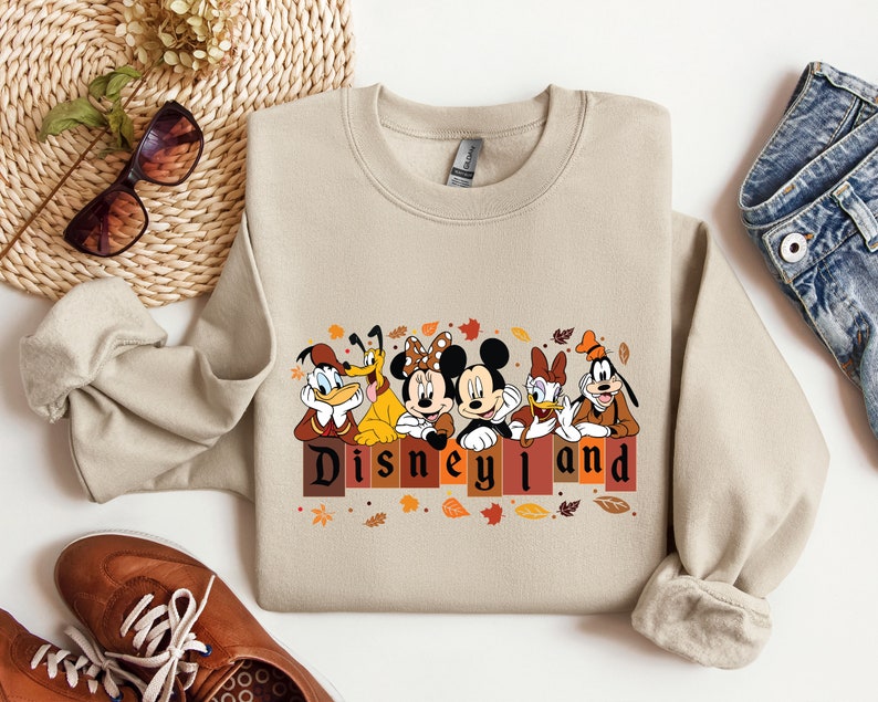 May include: A beige sweatshirt with a graphic of Mickey Mouse, Donald Duck, Goofy, and Pluto in front of a brick wall with the text "Disneyland" in brown letters. The characters are wearing fall-themed outfits and there are leaves scattered around them.