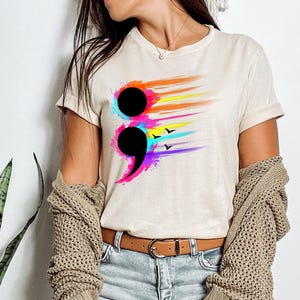 May include: Beige t-shirt featuring a black semicolon design with colorful brushstroke accents in orange, yellow, pink, purple, and blue. The design includes three small black bird silhouettes. The shirt has short sleeves and a crew neck.