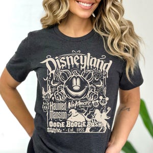 Puede incluir: Camiseta gris oscuro con un dise&ntilde;o crema. El dise&ntilde;o dice "Disneyland Haunted Mansion OOGIE BOOGIE BASH Est. 1955" e incluye una calabaza de Mickey Mouse, telara&ntilde;as y elementos de Halloween. Cuello redondo.