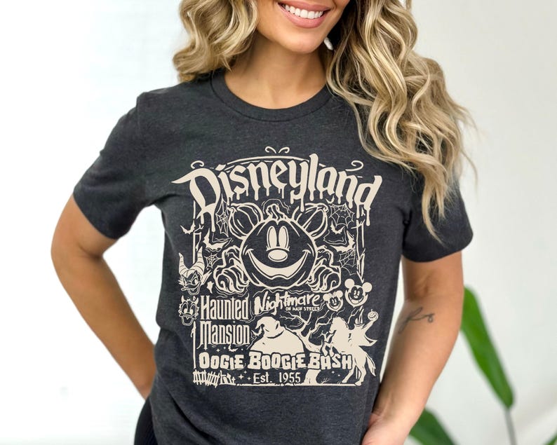 Puede incluir: Camiseta gris oscuro con un dise&ntilde;o crema. El dise&ntilde;o dice "Disneyland Haunted Mansion OOGIE BOOGIE BASH Est. 1955" e incluye una calabaza de Mickey Mouse, telara&ntilde;as y elementos de Halloween. Cuello redondo.