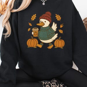 May include: Black sweatshirt featuring a cartoon duck wearing a knit hat and sweater, holding a coffee cup with a pumpkin design. Two pumpkins and fall leaves surround the duck. A cozy autumn-themed graphic.