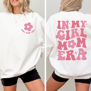 Puede incluir: Sudadera blanca con el texto "GIRL MOM ERA" y un gr&aacute;fico de flores rosas en la parte delantera. La parte trasera de la sudadera presenta la frase "IN MY GIRL MOM ERA" en rosa.