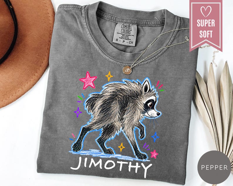 Puede incluir: Una camiseta gris carb&oacute;n con una ilustraci&oacute;n gr&aacute;fica de un mapache de pelaje esponjoso y marcas oscuras en los ojos. El dise&ntilde;o incluye estrellas coloridas de estilo dibujado a mano y el nombre JIMOTHY en letras may&uacute;sculas blancas debajo.