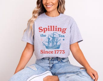 May include: A light gray t-shirt with the words "Spilling the Tea" in red and a blue ship graphic. The text "Since 1773" is below the ship. The shirt is worn by a person.