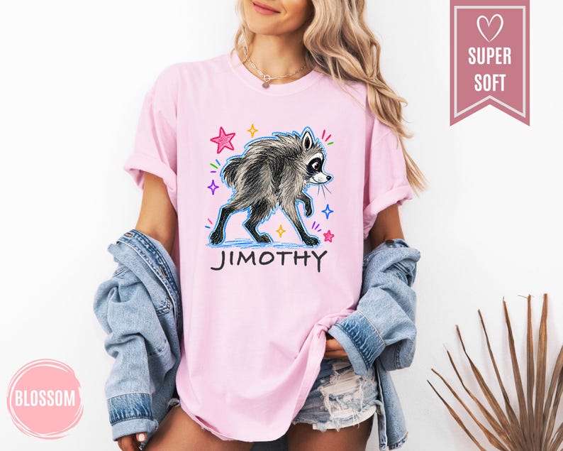 Puede incluir: Una camiseta de color rosa claro con un gr&aacute;fico de un mapache gris y el texto JIMOTHY debajo. El dise&ntilde;o incluye estrellas coloridas dibujadas a mano en rosa, azul y amarillo. La camiseta tiene mangas cortas y cuello redondo.