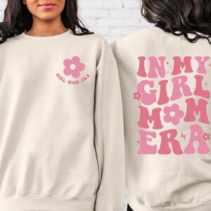 Puede incluir: Sudadera color crema con gr&aacute;ficos florales rosas. La parte delantera presenta una flor y el texto "GIRL MOM ERA". La parte trasera muestra "IN MY GIRL MOM ERA" en letras rosas.