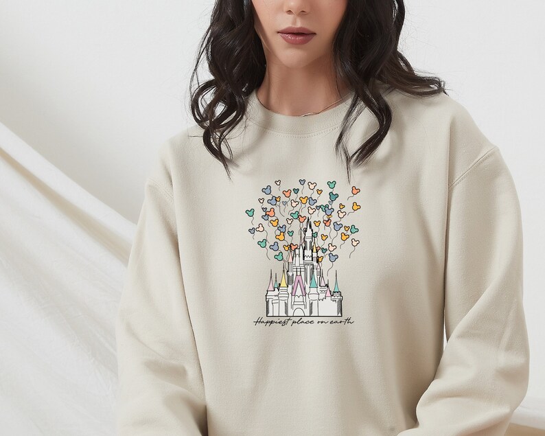 May include: A beige sweatshirt with a graphic of Cinderella's castle and colorful Mickey Mouse balloons. The text "Happiest place on earth" is printed below the castle.
