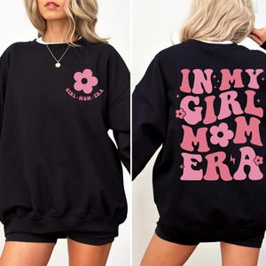 Puede incluir: Sudadera negra con ribete blanco, con un gr&aacute;fico de flor rosa y el texto "GIRL MOM ERA" en la parte delantera. La parte trasera muestra "IN MY GIRL MOM ERA" en rosa, con letras de estilo retro. Una prenda superior casual y c&oacute;moda.