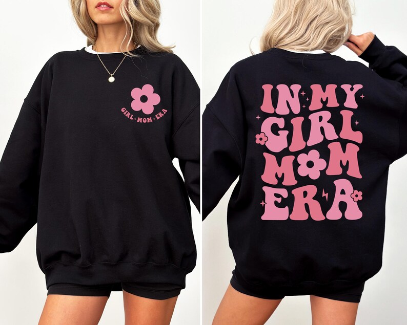 Puede incluir: Sudadera negra con ribete blanco, con un gr&aacute;fico de flor rosa y el texto "GIRL MOM ERA" en la parte delantera. La parte trasera muestra "IN MY GIRL MOM ERA" en rosa, con letras de estilo retro. Una prenda superior casual y c&oacute;moda.
