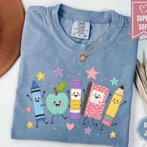 May include: A blue jean colored t-shirt featuring a graphic of a blue crayon, a green apple, a purple glue stick, a pink notebook, and a yellow pencil, all with smiling faces. The shirt has a Comfort Colors label and a pink banner reading SUPER SOFT.