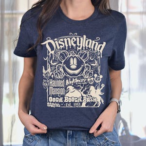 Puede incluir: Camiseta azul marino con un dise&ntilde;o gr&aacute;fico color crema. El dise&ntilde;o incluye la palabra "Disneyland" en una fuente estilizada, junto con una calabaza, Mickey Mouse y el texto "Haunted Nightmare" y "Oogie Boogie Bash Est. 1955".