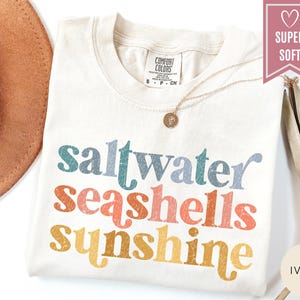May include: Ivory-colored t-shirt with the words "saltwater seashells sunshine" in colorful, retro-style lettering. The shirt has a crew neck and short sleeves. A gold necklace with a pendant is visible.