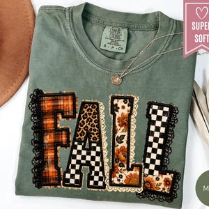 May include: A moss green short-sleeve t-shirt featuring the word FALL in large block letters. Each letter has a unique pattern including orange plaid, leopard print, black and white checkerboard, and floral prints, all outlined with black lace trim.