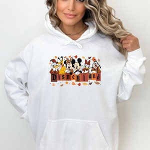 May include: White hoodie with a graphic of Mickey Mouse, Minnie Mouse, Goofy, Donald Duck, and Pluto in front of a sign that says "Disneyland". The graphic is surrounded by fall leaves.