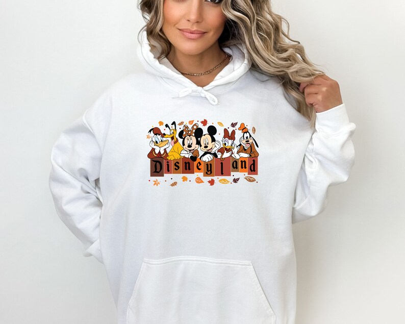 May include: White hoodie with a graphic of Mickey Mouse, Minnie Mouse, Goofy, Donald Duck, and Pluto in front of a sign that says "Disneyland". The graphic is surrounded by fall leaves.