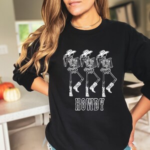 May include: Black sweatshirt with a white graphic of three skeletons wearing cowboy hats and boots. The text "HOWDY" is printed below the skeletons.