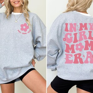 Puede incluir: Una sudadera gris claro con el texto "GIRL MOM ERA" y un gr&aacute;fico de flor rosa en la parte delantera. La parte trasera de la sudadera presenta el texto "IN MY GIRL MOM ERA" en rosa, con un estilo retro.