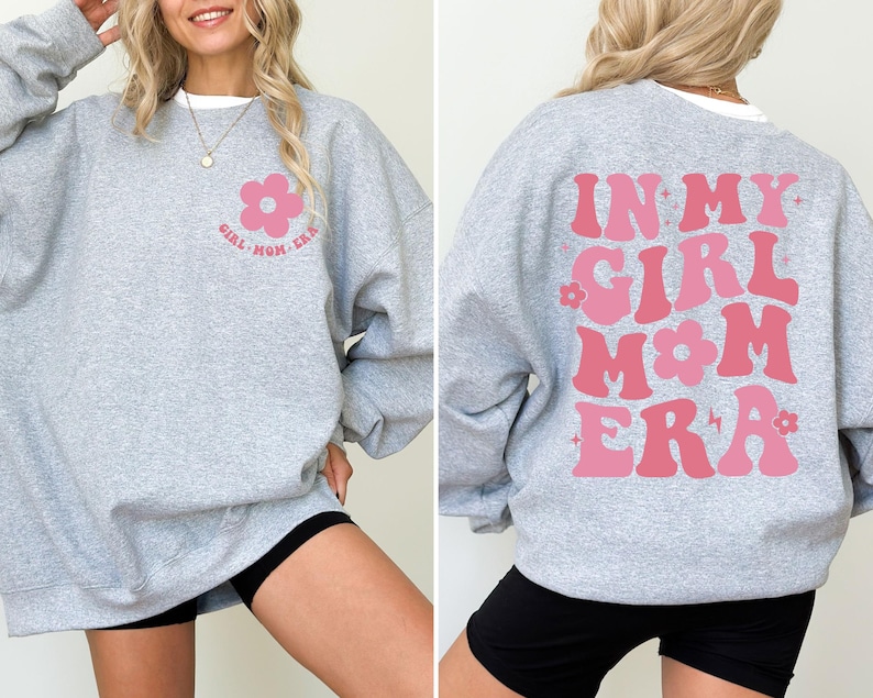 Puede incluir: Una sudadera gris claro con el texto "GIRL MOM ERA" y un gr&aacute;fico de flor rosa en la parte delantera. La parte trasera de la sudadera presenta el texto "IN MY GIRL MOM ERA" en rosa, con un estilo retro.