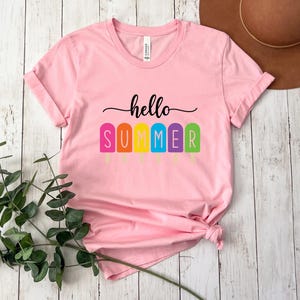 May include: A light pink t-shirt with the words "hello SUMMER" in black script and colorful popsicle graphics. The shirt is made of soft material and has rolled-up sleeves. The shirt is a summer-themed apparel item.