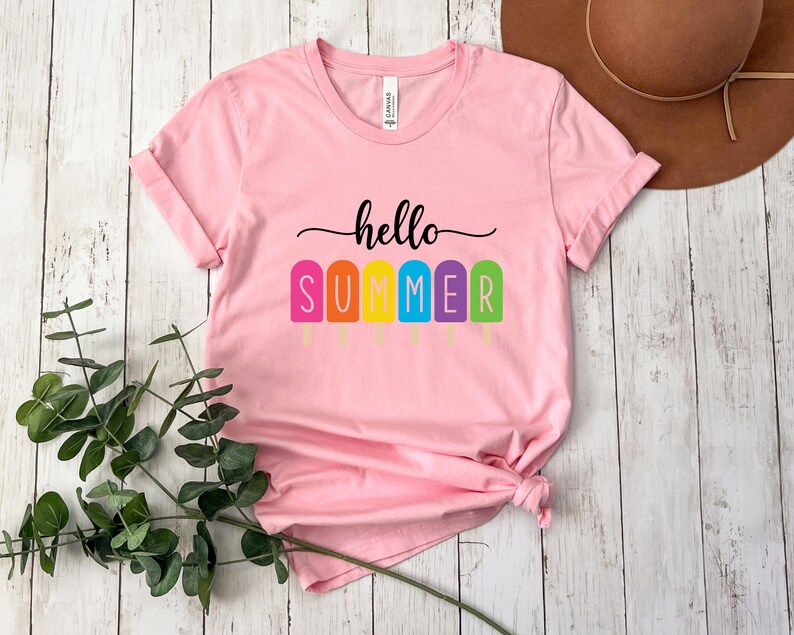 May include: A light pink t-shirt with the words "hello SUMMER" in black script and colorful popsicle graphics. The shirt is made of soft material and has rolled-up sleeves. The shirt is a summer-themed apparel item.