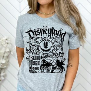 Puede incluir: Camiseta gris claro con un dise&ntilde;o gr&aacute;fico negro. El dise&ntilde;o presenta el texto "Disneyland", "Haunted Mansion Oogie Boogie Bash Est. 1955", una calabaza sonriente, Mickey Mouse y otros personajes tem&aacute;ticos de Halloween.