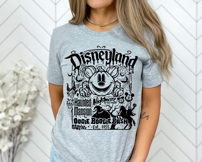 Puede incluir: Camiseta gris claro con un dise&ntilde;o gr&aacute;fico negro. El dise&ntilde;o presenta el texto "Disneyland", "Haunted Mansion Oogie Boogie Bash Est. 1955", una calabaza sonriente, Mickey Mouse y otros personajes tem&aacute;ticos de Halloween.