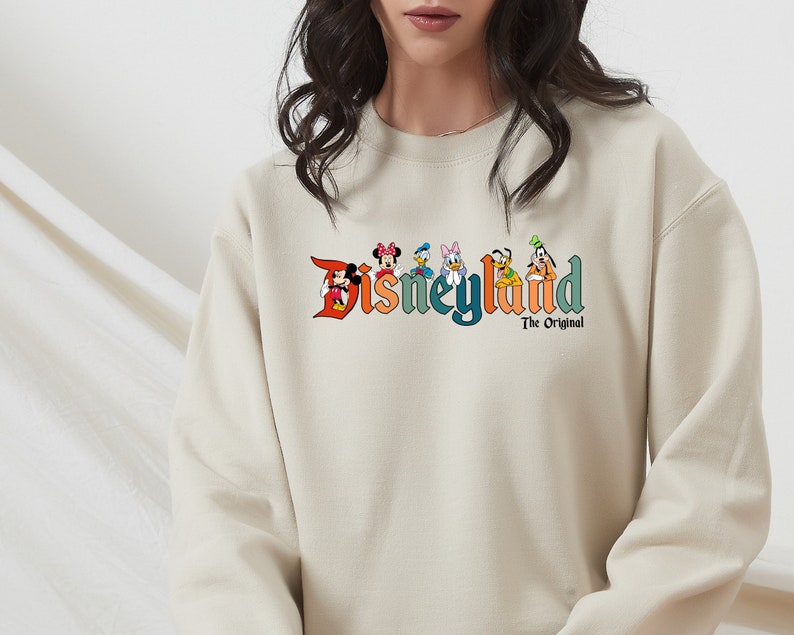 Puede incluir: Una sudadera beige con un gr&aacute;fico colorido de personajes de Disney y el texto "Disneyland The Original".