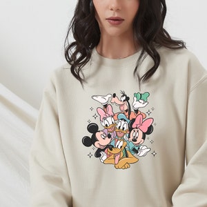 K&ouml;nnte beinhalten: Ein beiger Pullover mit einem bunten Grafikdruck von Mickey Mouse, Minnie Mouse, Donald Duck, Goofy und Pluto. Die Figuren sind in einer verspielten, gestapelten Formation angeordnet.