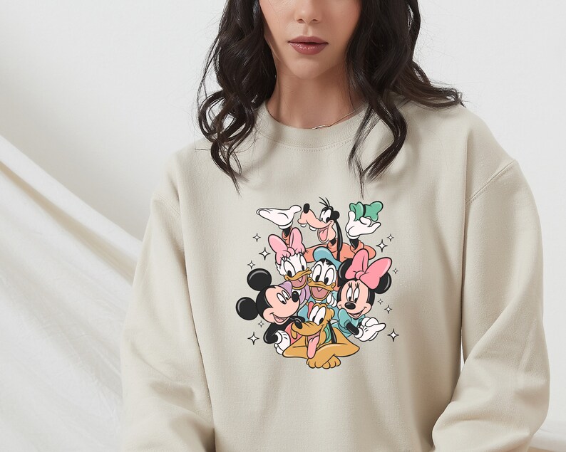 K&ouml;nnte beinhalten: Ein beiger Pullover mit einem bunten Grafikdruck von Mickey Mouse, Minnie Mouse, Donald Duck, Goofy und Pluto. Die Figuren sind in einer verspielten, gestapelten Formation angeordnet.