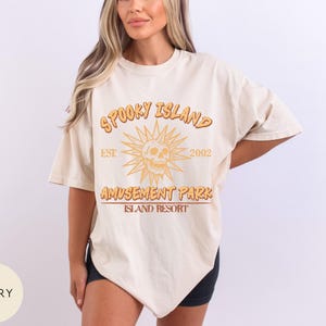 May include: Ivory-colored t-shirt with the text "Spooky Island Amusement Park Island Resort" in orange and brown. The design includes a skull with sun rays and the year "2002". The shirt has short sleeves and a relaxed fit.