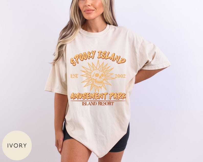 May include: Ivory-colored t-shirt with the text "Spooky Island Amusement Park Island Resort" in orange and brown. The design includes a skull with sun rays and the year "2002". The shirt has short sleeves and a relaxed fit.