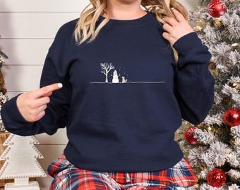 May include: Navy blue sweatshirt featuring a minimalist winter scene. The design includes a snowman, a dog, and a bare tree under a sprinkling of snow. The sweatshirt is a comfortable, casual style, perfect for the holiday season.