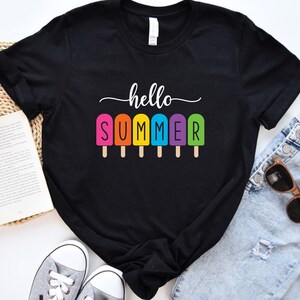May include: Black t-shirt with the words "hello SUMMER" in white script above a row of colorful popsicle graphics. The popsicles are pink, orange, yellow, blue, and green. The shirt is paired with blue jeans, sunglasses, and sneakers.