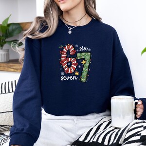 May include: Navy blue sweatshirt with a festive design featuring the numbers "67" decorated with candy cane stripes and green lights. The word "six" and "seven" are written above and below the numbers. A white mug is held in the hand.