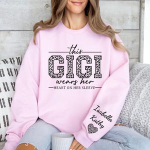 May include: Pink sweatshirt with the text "This Gigi wears her heart on her sleeve" in black with a leopard print design. The sleeve has the names "Isabella Kathy" and a small heart.