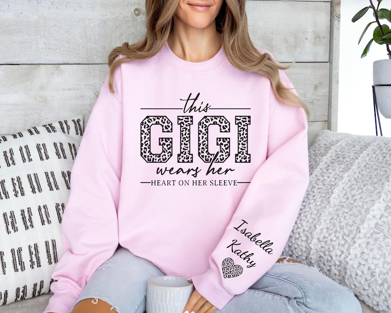 May include: Pink sweatshirt with the text "This Gigi wears her heart on her sleeve" in black with a leopard print design. The sleeve has the names "Isabella Kathy" and a small heart.