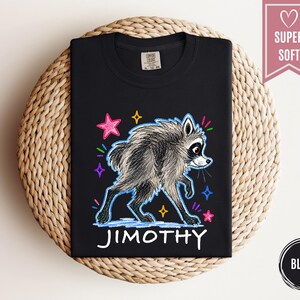 Puede incluir: Una camiseta negra con una ilustraci&oacute;n gr&aacute;fica de un mapache gris con un aspecto texturizado y de boceto. El dise&ntilde;o incluye estrellas coloridas de estilo ne&oacute;n y el nombre JIMOTHY impreso en letras may&uacute;sculas blancas debajo del mapache.