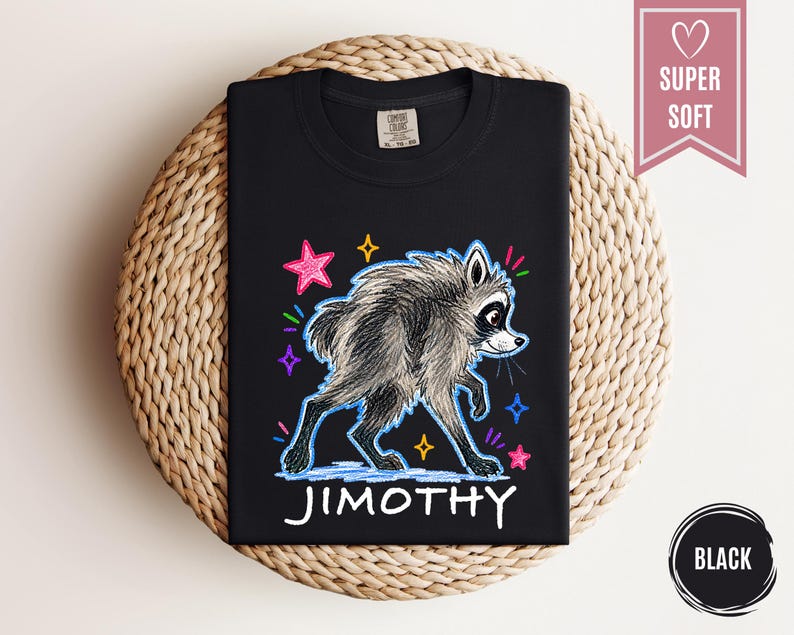 Puede incluir: Una camiseta negra con una ilustraci&oacute;n gr&aacute;fica de un mapache gris con un aspecto texturizado y de boceto. El dise&ntilde;o incluye estrellas coloridas de estilo ne&oacute;n y el nombre JIMOTHY impreso en letras may&uacute;sculas blancas debajo del mapache.