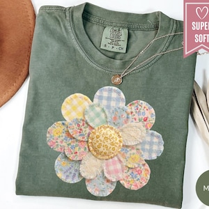 May include: A moss green t-shirt with a large, appliqued flower design. The flower is made of various patterned fabric patches in pastel colors. A delicate silver necklace with a pendant rests on the shirt. The shirt has a Comfort Colors tag.