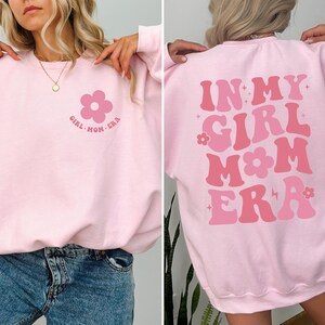 Puede incluir: Una sudadera rosa claro con el texto "IN MY GIRL MOM ERA" en tonos rosas en la espalda. La parte delantera presenta un gr&aacute;fico de flores y el texto "GIRL MOM ERA".