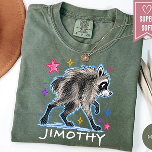 Puede incluir: Una camiseta verde oliva con una ilustraci&oacute;n gr&aacute;fica de un mapache con pelaje blanco y negro. El dise&ntilde;o incluye estrellas y destellos coloridos dibujados a mano en rosa, amarillo y morado, con el nombre JIMOTHY impreso en blanco debajo.