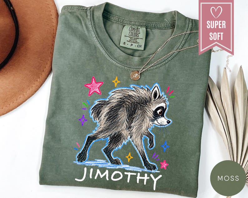 Puede incluir: Una camiseta verde oliva con una ilustraci&oacute;n gr&aacute;fica de un mapache con pelaje blanco y negro. El dise&ntilde;o incluye estrellas y destellos coloridos dibujados a mano en rosa, amarillo y morado, con el nombre JIMOTHY impreso en blanco debajo.