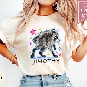 Puede incluir: Una camiseta de color marfil con un gr&aacute;fico de un mapache gris con contorno azul, rodeado de estrellas de colores y el texto "JIMOTHY" en negro. Una banda rosa en la esquina superior derecha contiene un icono de coraz&oacute;n y el texto "SUPER SOFT".