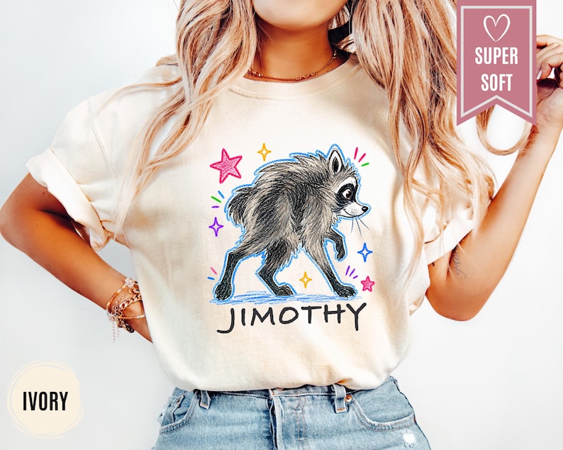 Puede incluir: Una camiseta de color marfil con un gr&aacute;fico de un mapache gris con contorno azul, rodeado de estrellas de colores y el texto "JIMOTHY" en negro. Una banda rosa en la esquina superior derecha contiene un icono de coraz&oacute;n y el texto "SUPER SOFT".