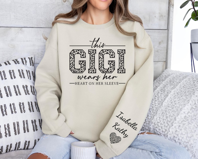 May include: A beige sweatshirt with the text "This Gigi wears her heart on her sleeve" in black lettering. The word "Gigi" is decorated with a leopard print pattern. The sleeve has the names "Isabella Kathy" and a small heart.