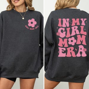 Puede incluir: Sudadera gris oscuro con dise&ntilde;o floral y texto en rosa. La parte delantera presenta una flor rosa y las palabras "GIRL MOM ERA". La parte trasera muestra "IN MY GIRL MOM ERA" en rosa, letras retro.